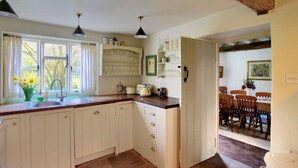 Una cocina con armarios blancos, un fregadero y una ventana con cortinas. Hay un comedor con sillas de madera y una mesa.