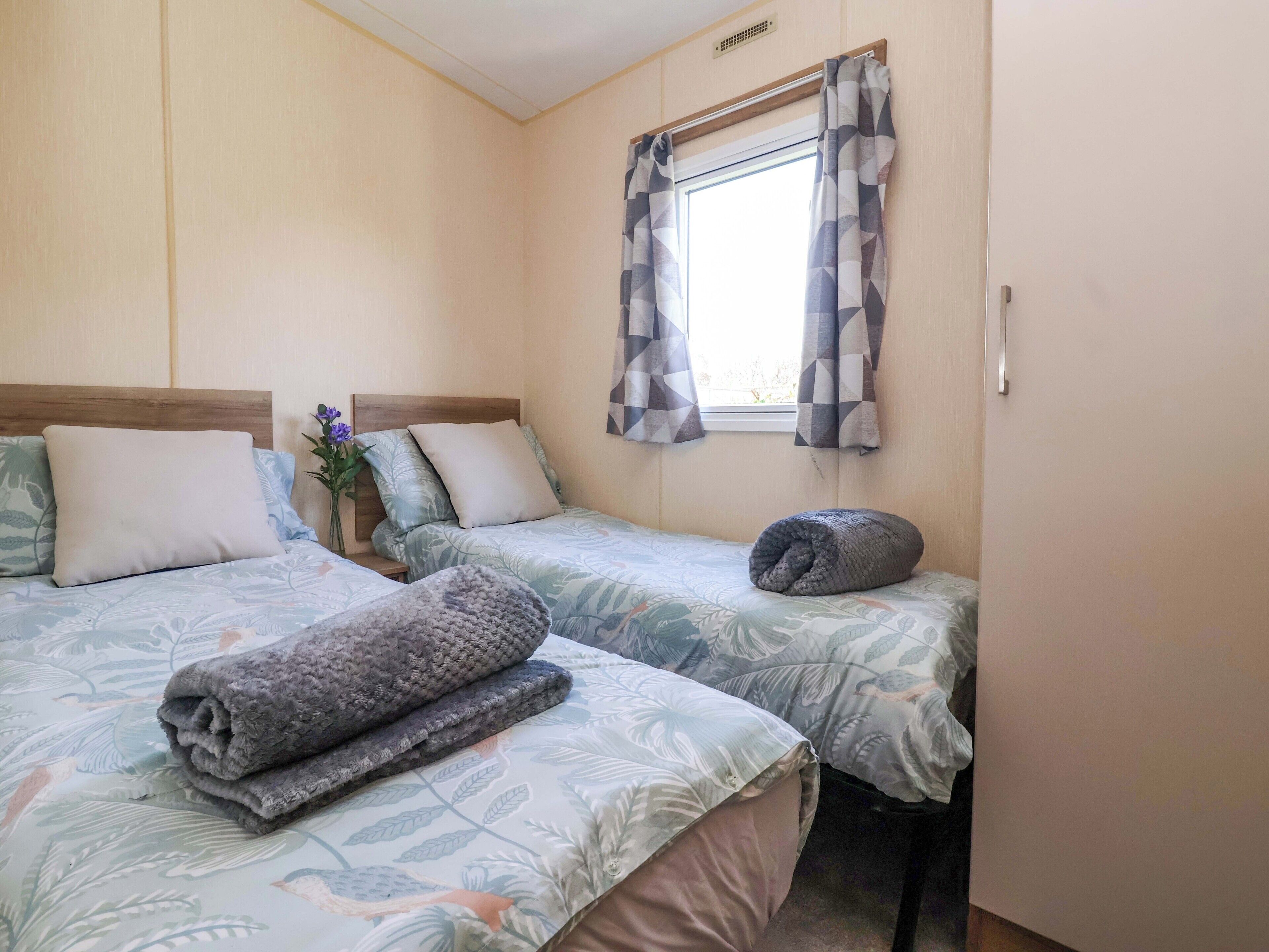 Une chambre avec deux lits, une fenêtre avec des rideaux, une table de chevet ornée d’un vase de fleurs et un radiateur fixé au mur.