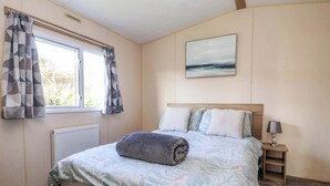 Un dormitorio con cama, ventana con cortinas, radiador, mesita de noche con lámpara y un cuadro en la pared.