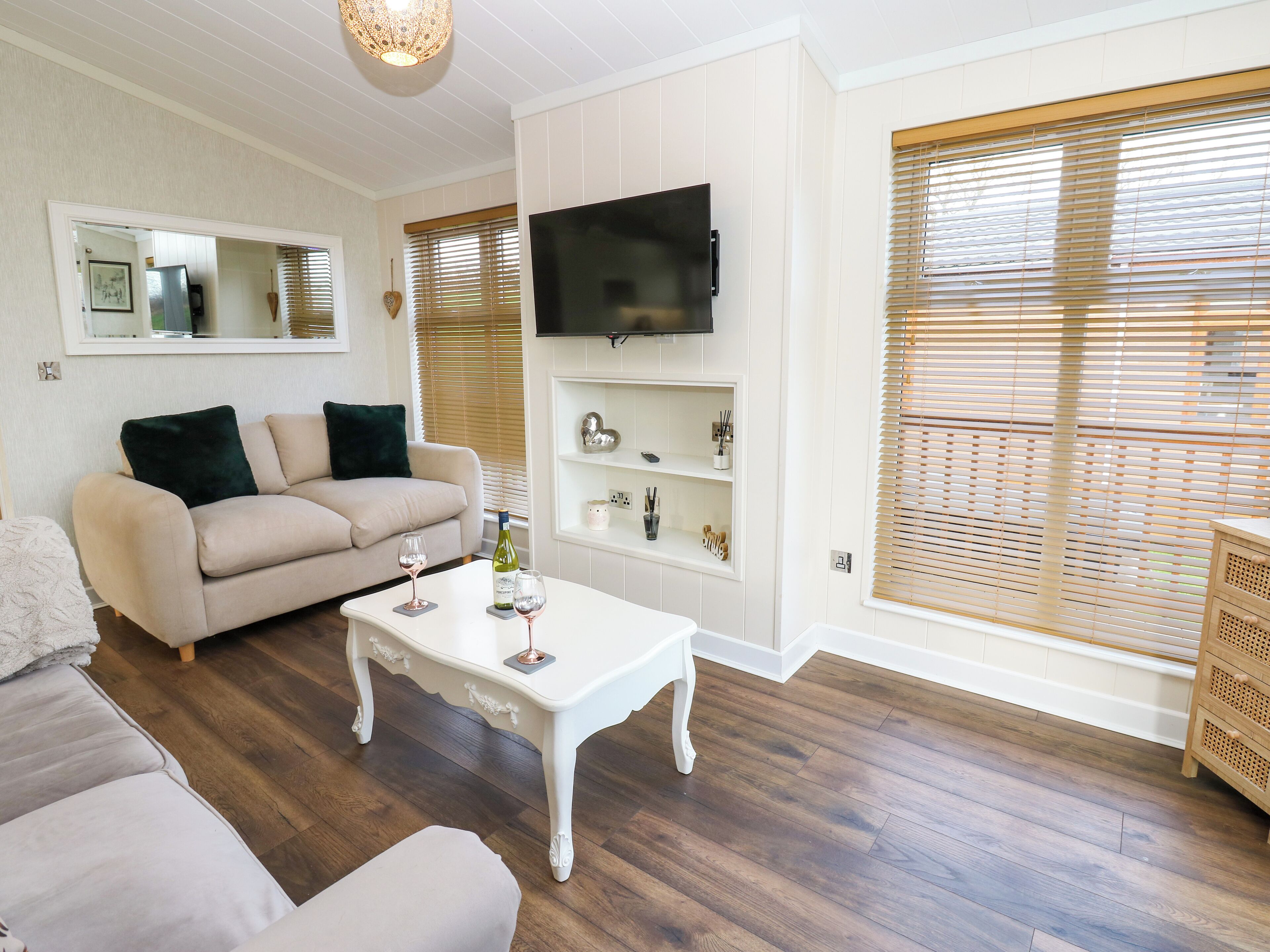 A living room with a beige sofa, a white coffee table with wine glasses, a flat-screen TV mounted on the wall, and wooden blinds on the windows.