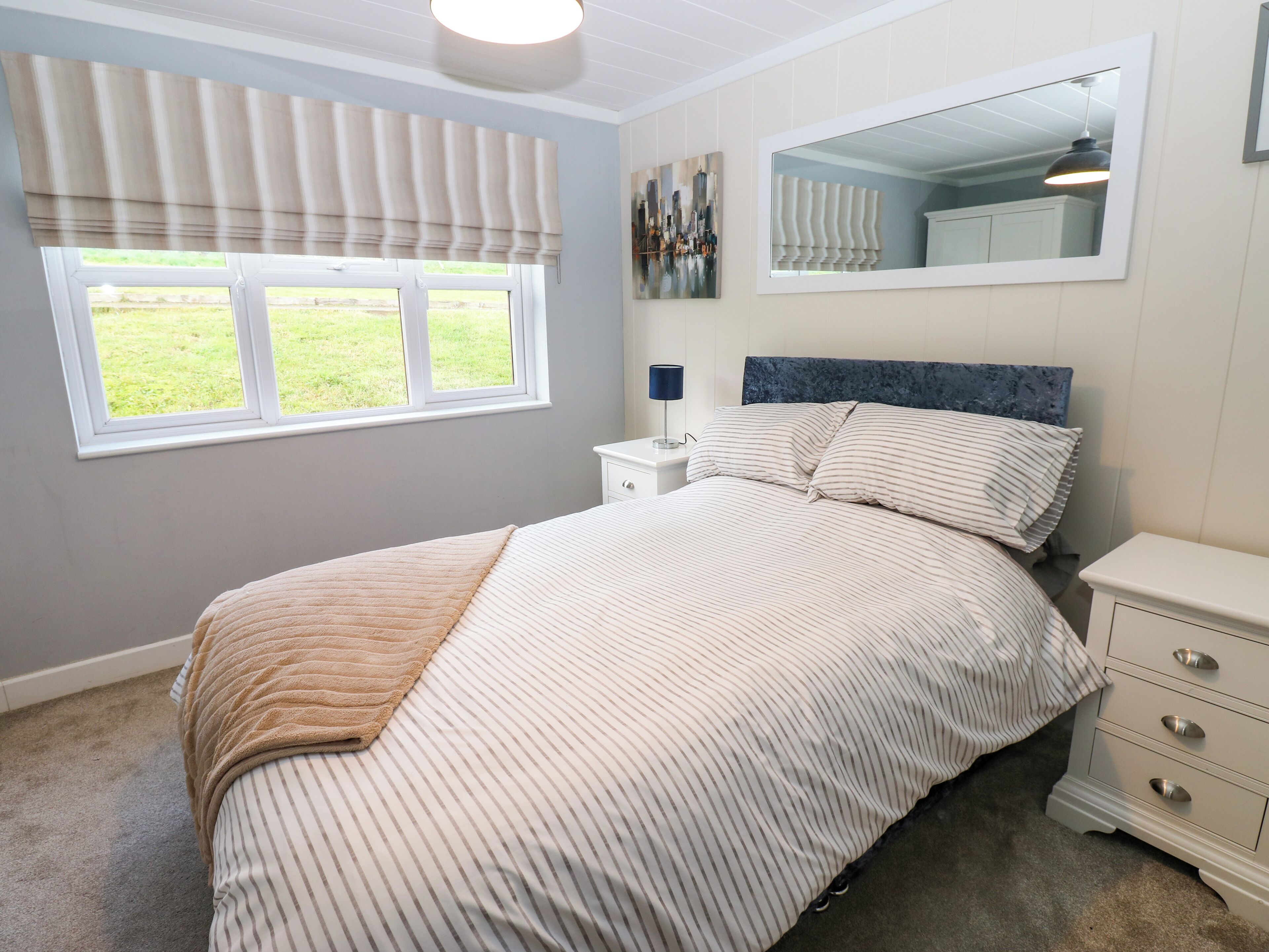 A bedroom with a bed, a nightstand, a dresser, a window with blinds, a mirror, and a painting on the wall.