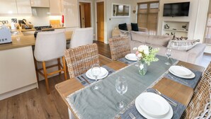 Une cuisine moderne avec des meubles blancs, un coin repas avec une table dressée pour deux personnes, et un salon avec un canapé et un téléviseur.
