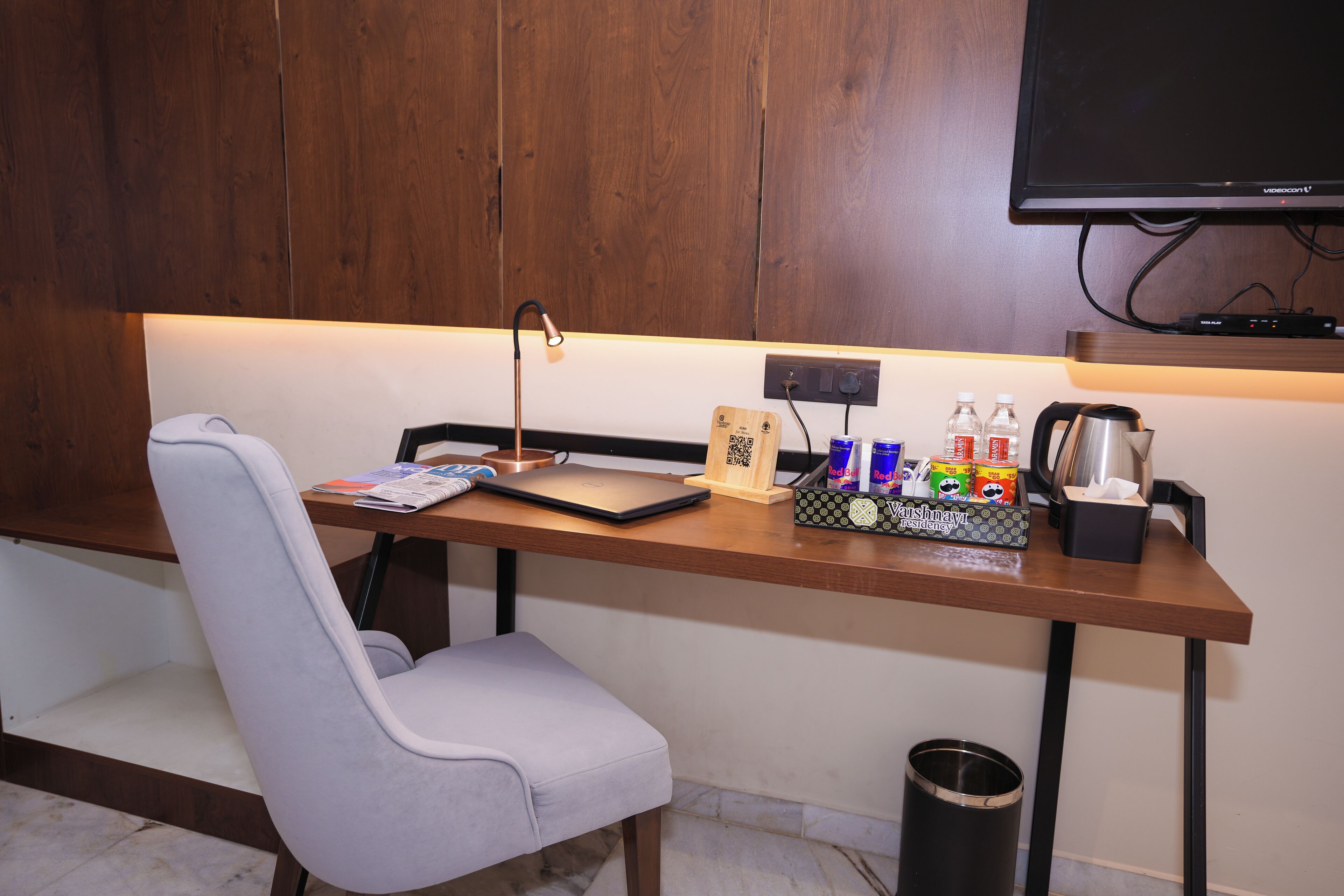 Une chambre d’hôtel moderne avec un bureau en bois, un ordinateur portable, une chaise, une petite poubelle et un téléviseur fixé au mur.