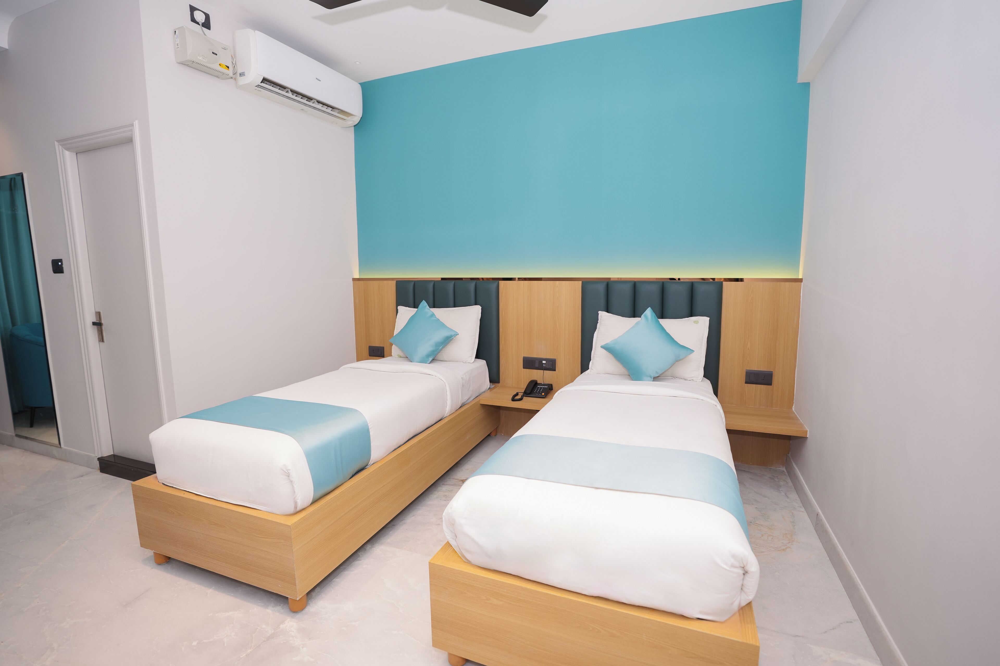 A hotel room with two beds, each with a wooden frame and blue bedding. There is an air conditioning unit on the wall, and a small bedside table with a phone on it.