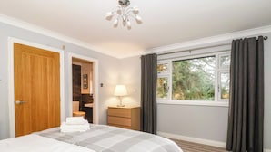 A hotel room with a bed, a nightstand, a lamp, a window with curtains, and a door. - The Pines (Skipton)