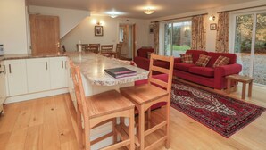 Een ruime woonkamer met een rode bank, houten stoelen, een keuken met een marmeren aanrecht en een groot raam met gordijnen.