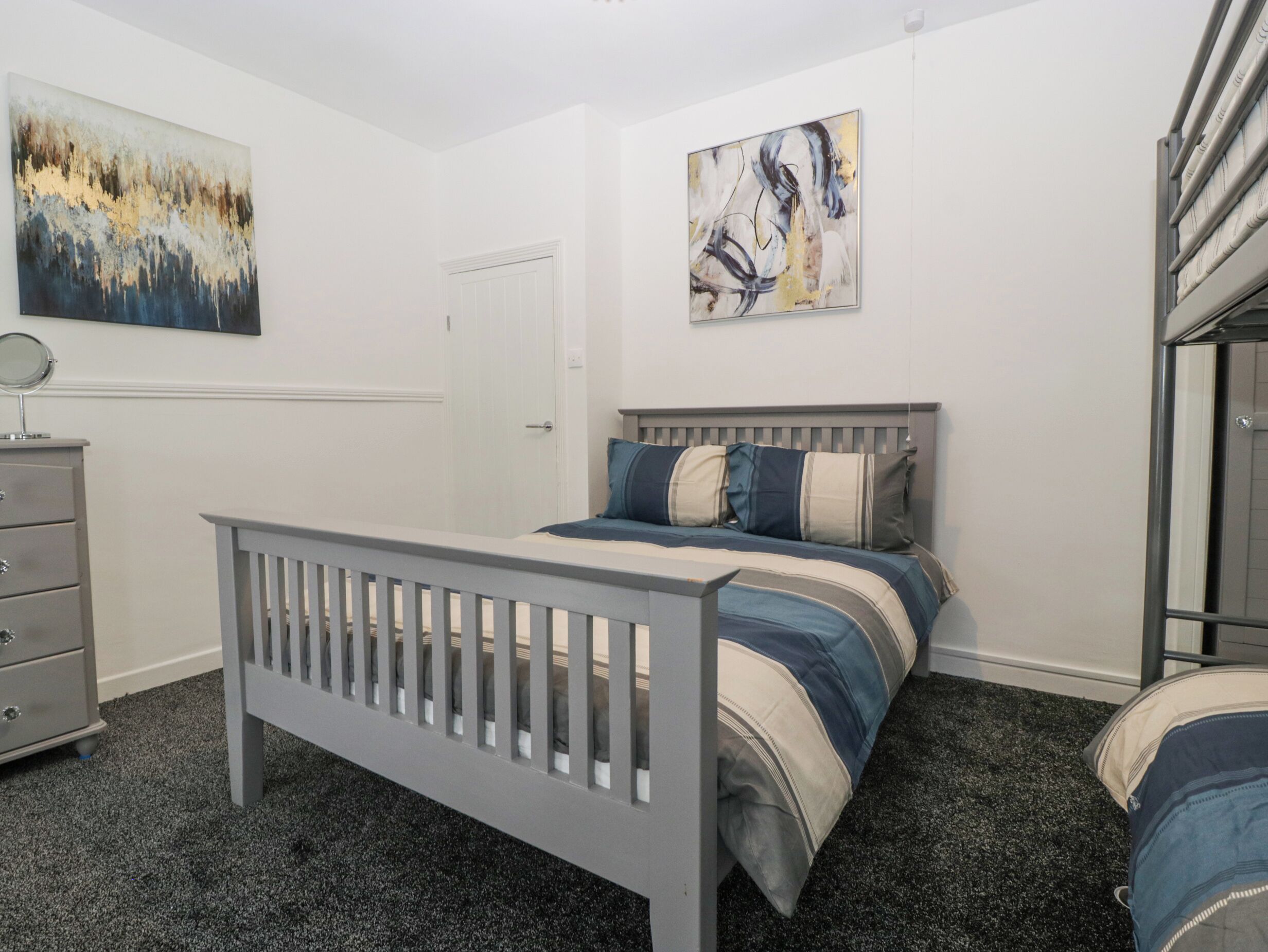 Un dormitorio con una cama gris, un aparador y dos cuadros abstractos en las paredes.
