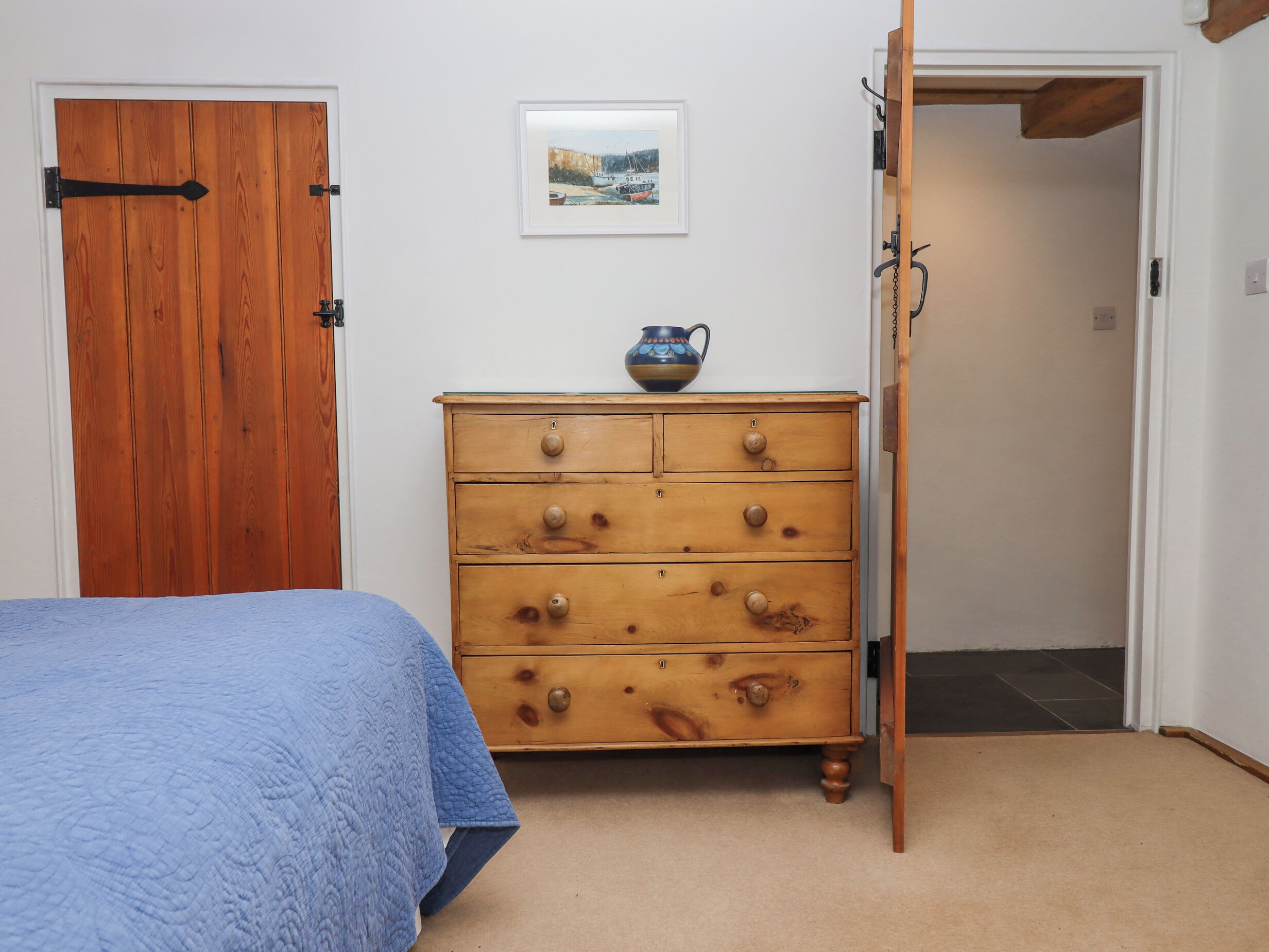 Un dormitorio con un aparador de madera, una cama con colcha azul, un cuadro enmarcado en la pared y una puerta que da a otra habitación.