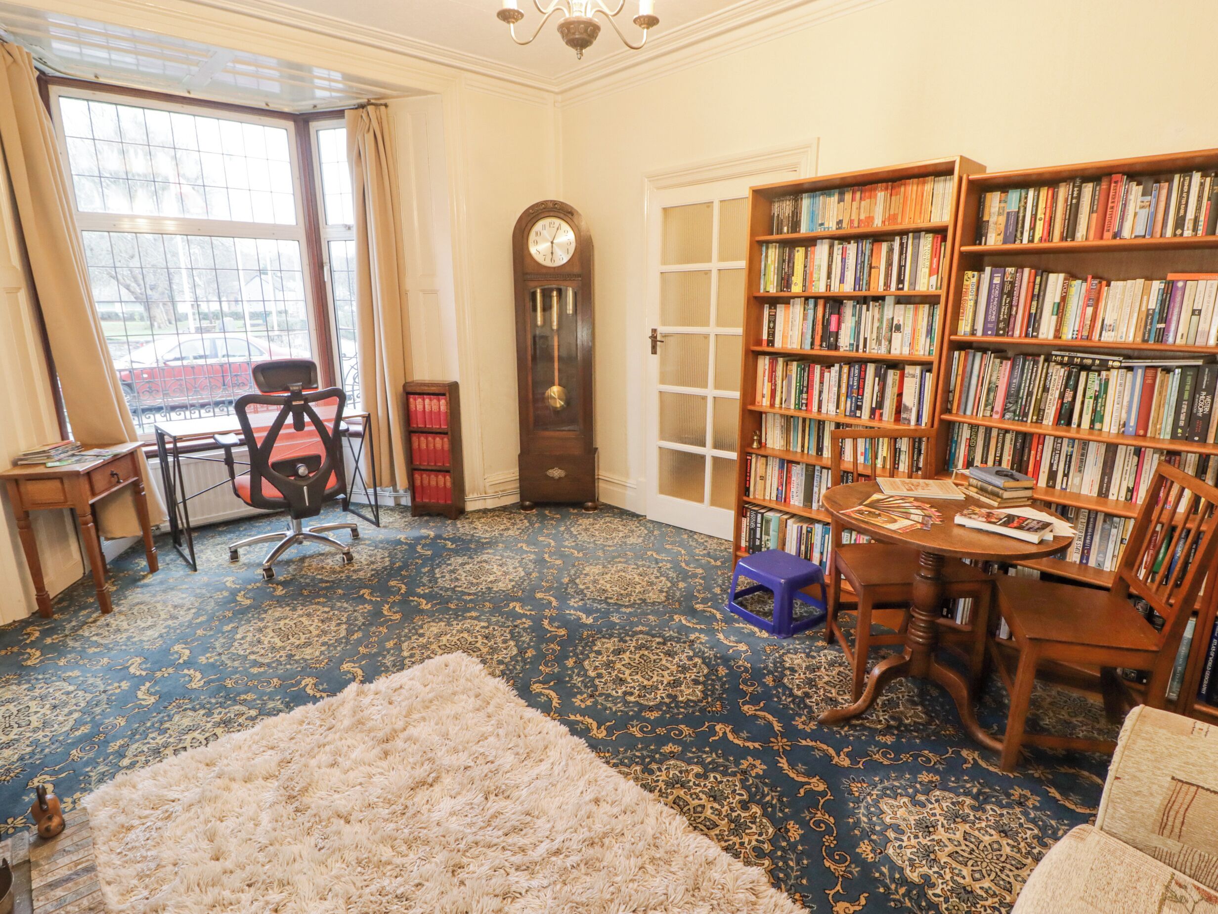 A library with bookshelves, a desk, a chair, a clock, and a large window.
