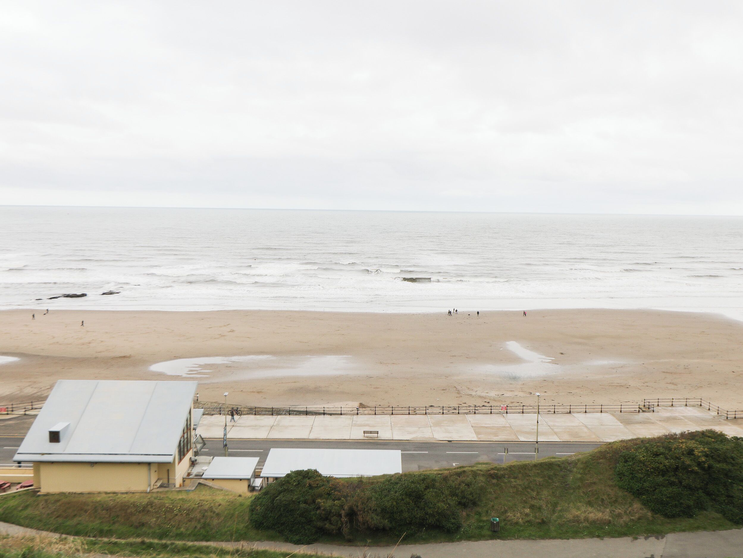 Een strand met een zandige kust, enkele mensen die wandelen en een gebouw met een plat dak op de voorgrond.