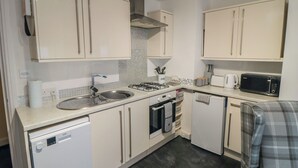 A compact kitchen with white cabinets, a dishwasher, a stove, a microwave, and a small seating area. - Kittiwake (Scarborough)