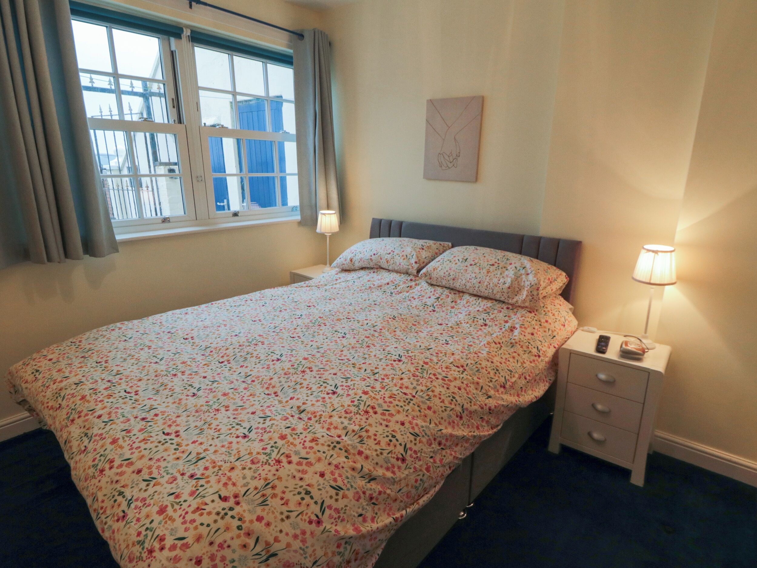 A bedroom with a floral bedspread, a bedside table with a lamp, and a window with curtains.