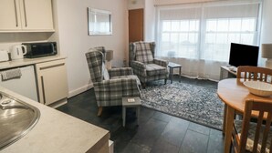 A compact living space with a kitchenette, two plaid armchairs, a small table, a TV, and a window with sheer curtains. - Kittiwake (Scarborough)