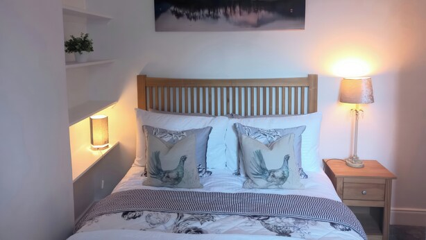 A bedroom with a wooden headboard, a bed with striped bedding, two pillows with a rooster design, a bedside lamp, and a wall-mounted painting of a mountain landscape. - Lismore (Crieff)