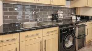 A modern kitchen with light wood cabinets, a black tiled backsplash, stainless steel appliances, and a built-in washer and dryer. - Lismore (Crieff)