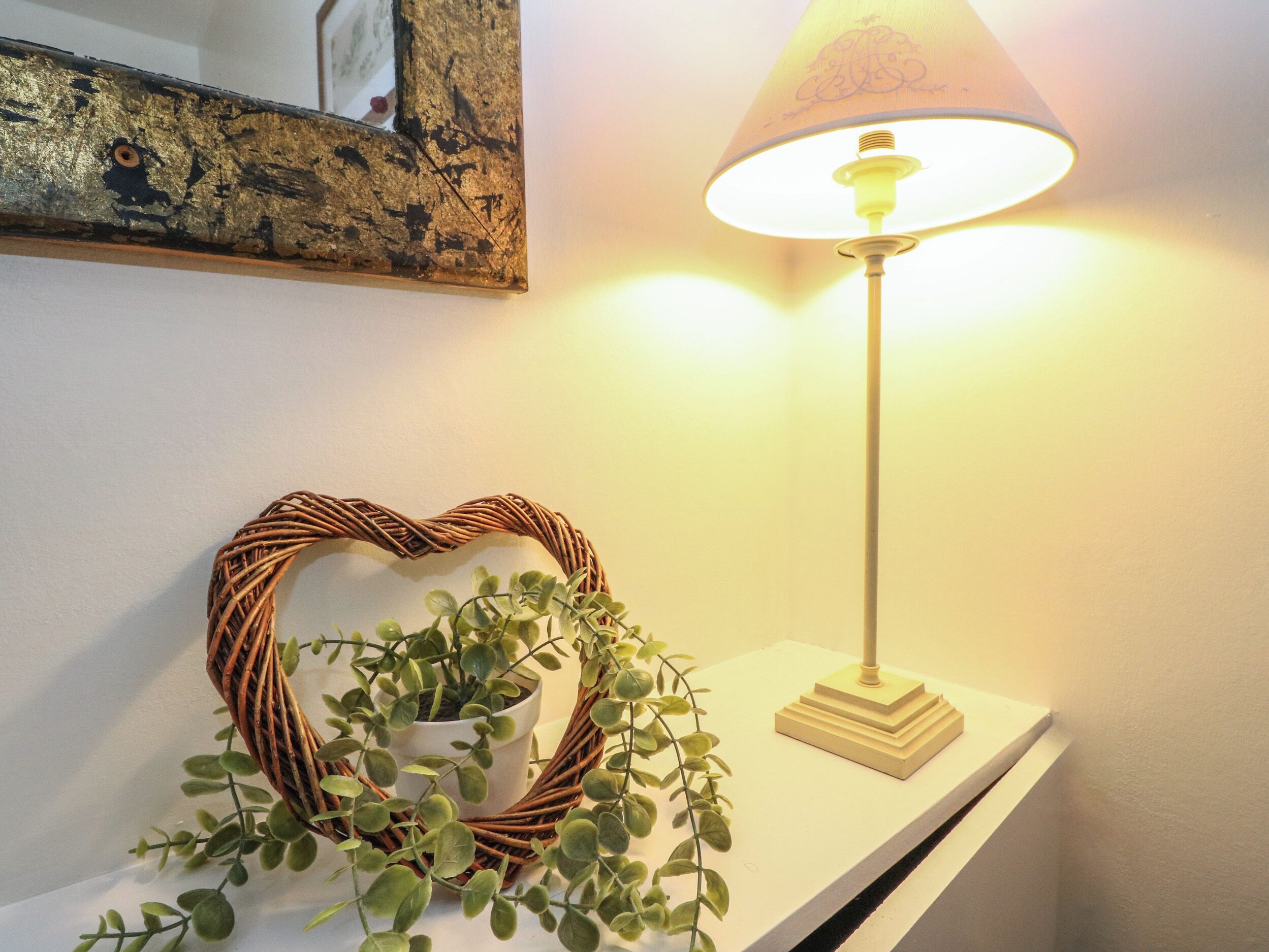 Un miroir mural avec un cadre vieilli, une décoration en forme de cœur faite d’osier dans laquelle est placée une plante en pot, et une lampe de table avec un abat-jour blanc.