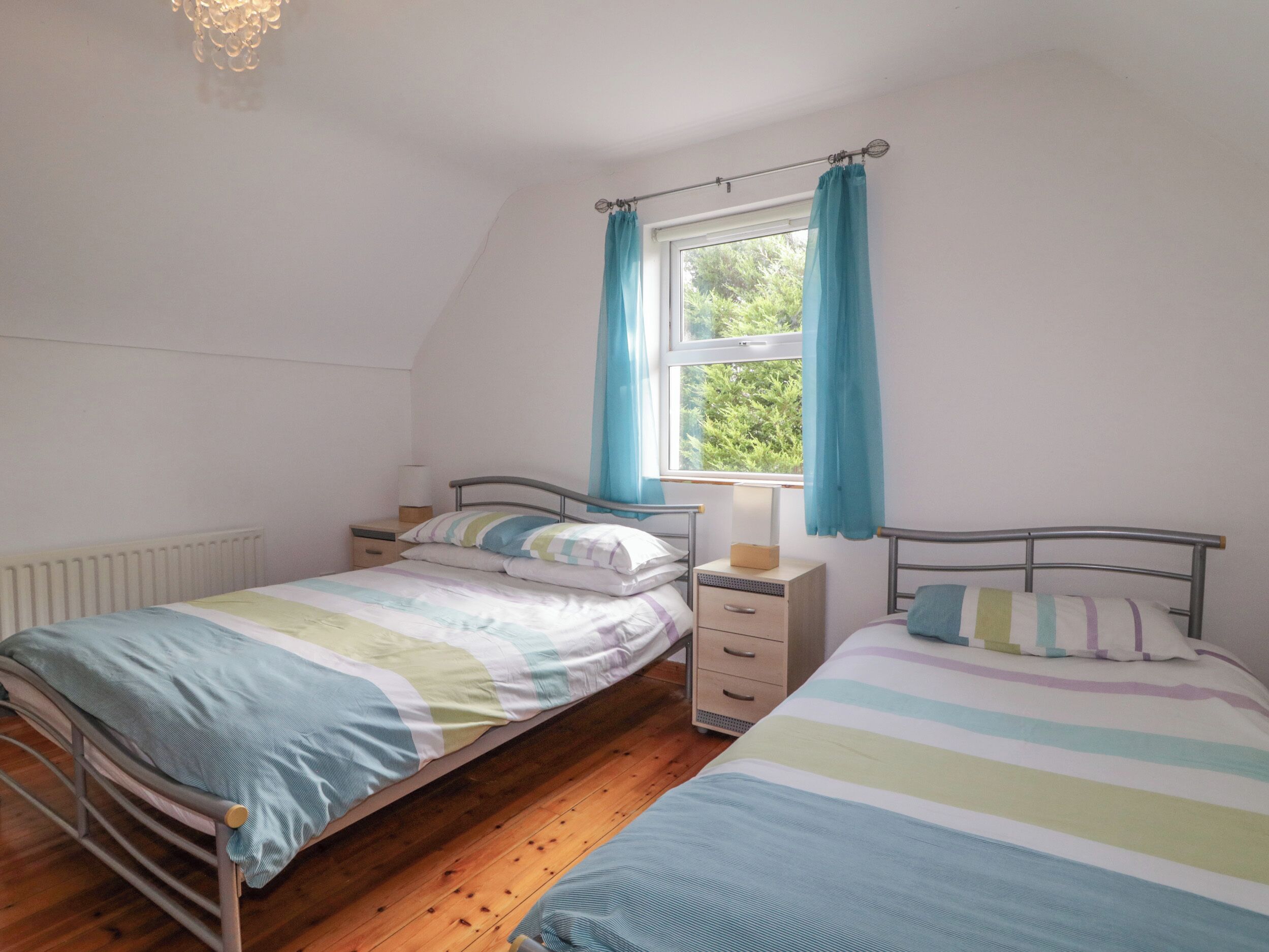 Un dormitorio con dos camas individuales, una ventana con cortinas, una mesita de noche con lámpara y un radiador.