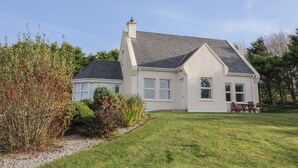 A two-story house with a sloped roof, a front porch, and a small garden area with shrubs and a lawn. - Horn Head View (Carrigart)