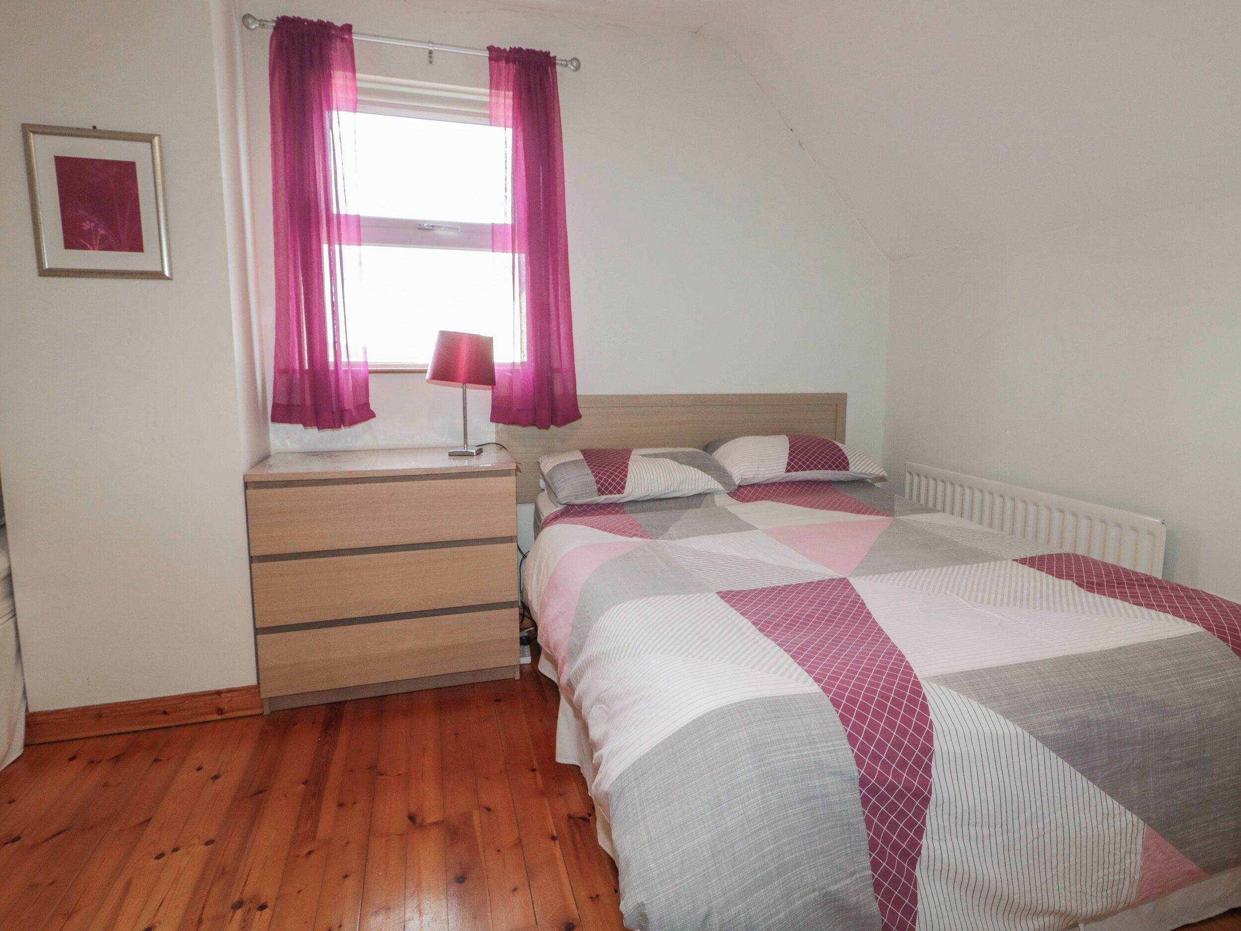 A bedroom with a bed, a nightstand, a lamp, a window with curtains, and a radiator.