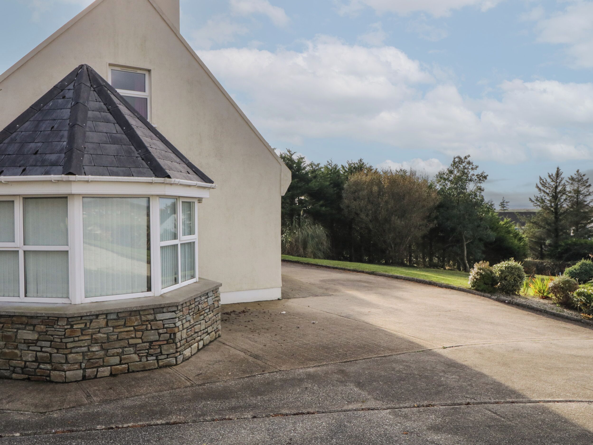A house with a stone base and a dark roof, a bay window, and a driveway leading to a grassy area with trees.