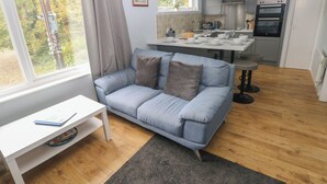 A modern living room with a grey sofa, a small white table, and a kitchen area with a dining table set for four. - Kittiwake, Apartment 22 (Haverfordwest)