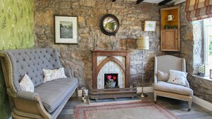 A cozy room with a stone wall, a fireplace, a sofa, a chair, and a window with curtains. - Goleufryn (Beaumaris)