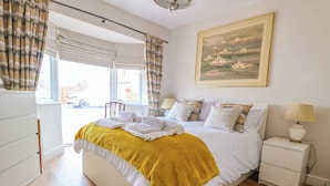 A bedroom with a large bed, a bedside table with a lamp, a window with curtains, and a painting on the wall. - Brimrod (Holywell)