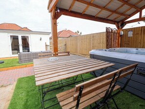 A covered patio area with a wooden table and benches, a hot tub, and a fenced backyard. - Brimrod (Holywell)