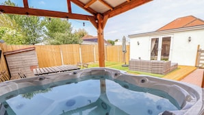 A hot tub in a covered outdoor area with a wooden deck and a white building in the background. - Brimrod (Holywell)