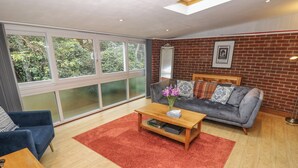 A modern living room with a grey sofa, blue armchairs, a wooden coffee table, and a red rug. Large windows offer a view of greenery outside. - Medina by The Solent (Cowes)