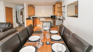 Un comedor con una mesa de madera dispuesta para seis personas, rodeada de sillas de cuero. Se ve una cocina con armarios de madera y una encimera al fondo.