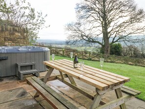 Une table de pique-nique en bois, avec deux verres à vin et une bouteille, placée devant un mur de pierre, avec en arrière-plan un champ verdoyant et des arbres.