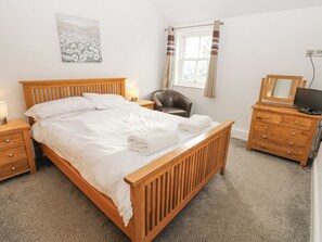 Un lit bien fait, avec du linge de lit blanc, une table de chevet en bois avec une lampe, une commode en bois avec un miroir et un téléviseur, et une chaise dans une pièce aux murs blancs et ornée d’un tableau.