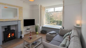 A cozy living room with a fireplace, a flat-screen TV, a comfortable sofa, and a window with a view of trees. - Trackside (Whitby)
