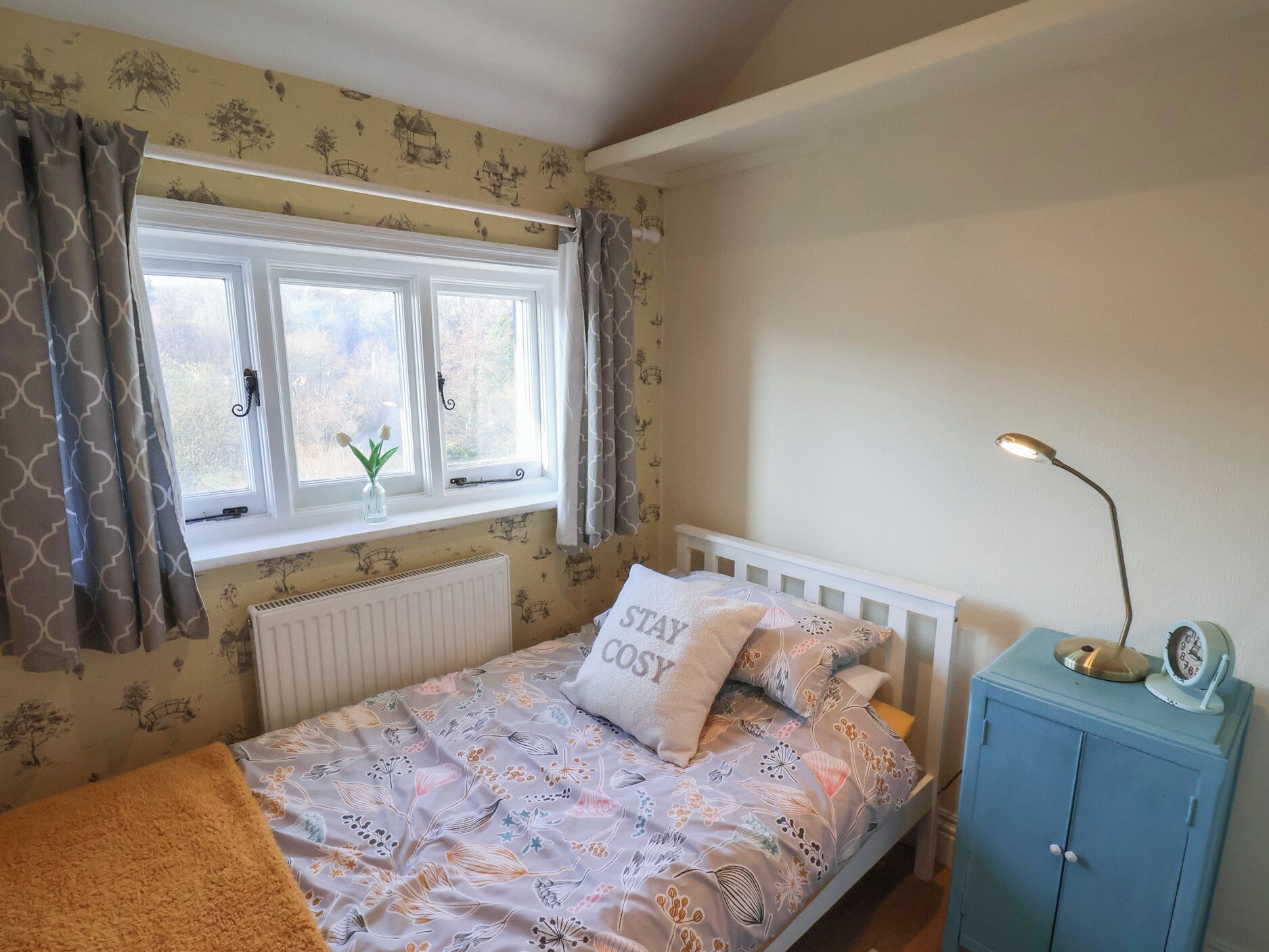 Una camera da letto con un letto, una finestra con tende, un termosifone, un comodino con una lampada e una sveglia.