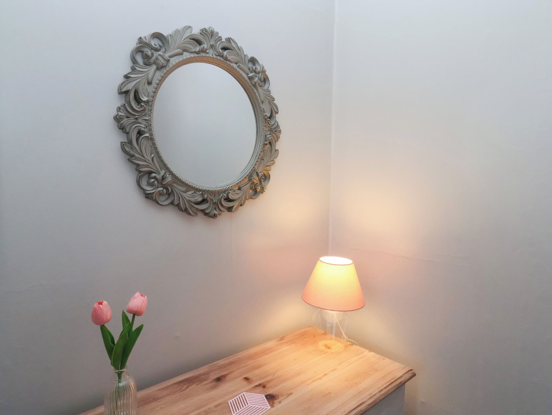 Et spejl med et detaljeret ramme på en væg, et trænatbord med en lampe, og en vase med lyserøde tulipaner.