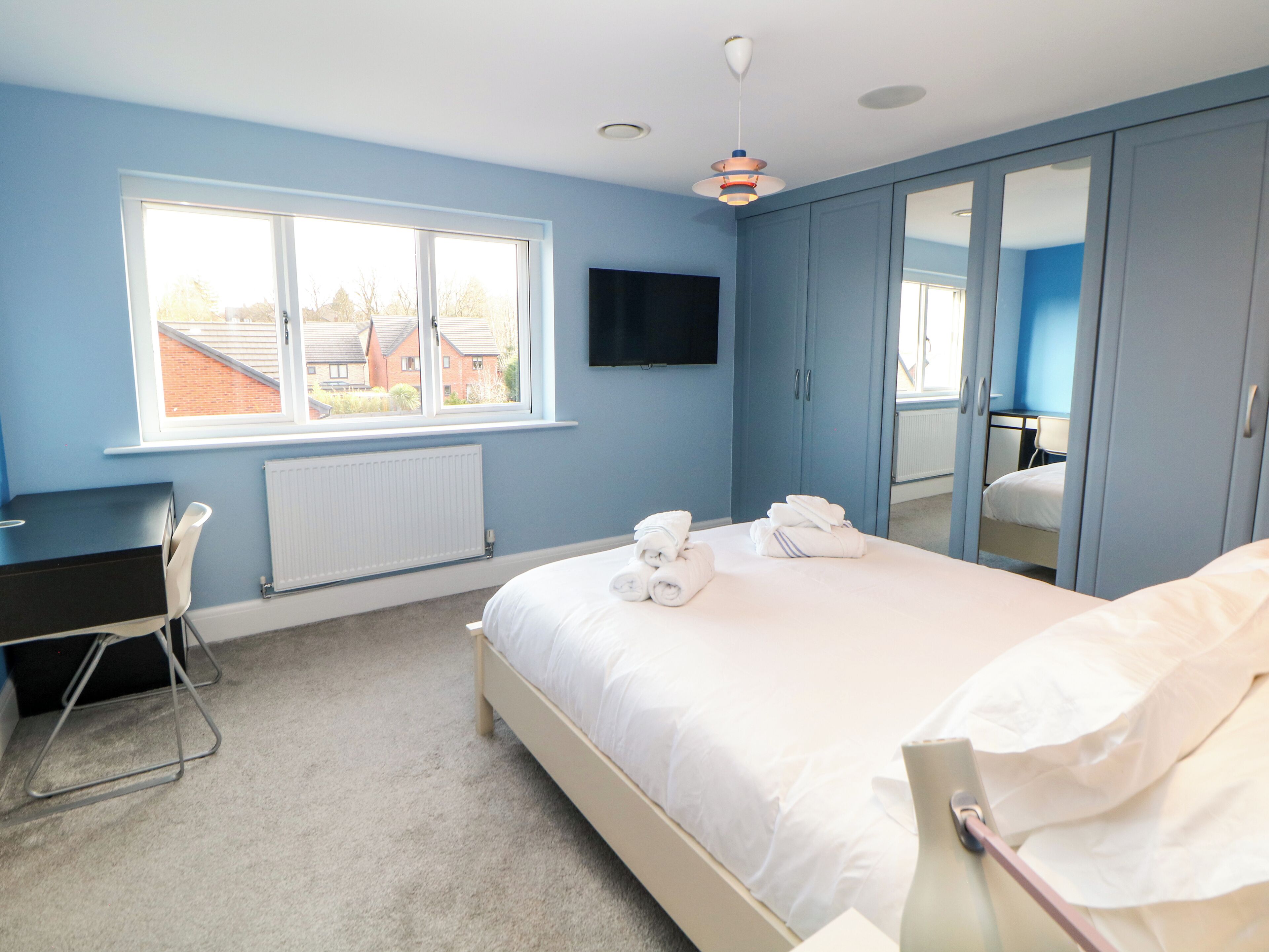 A bedroom with a bed, a desk, a chair, a TV, and a window with a view of buildings.
