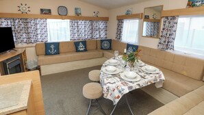 Un interior de autocaravana acogedor con comedor para cuatro, un sofá con cojines de temática náutica y un televisor sobre un soporte de madera.