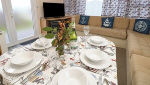 Un comedor con una mesa preparada para cuatro, un sofá, un televisor y una ventana con cortinas estampadas.