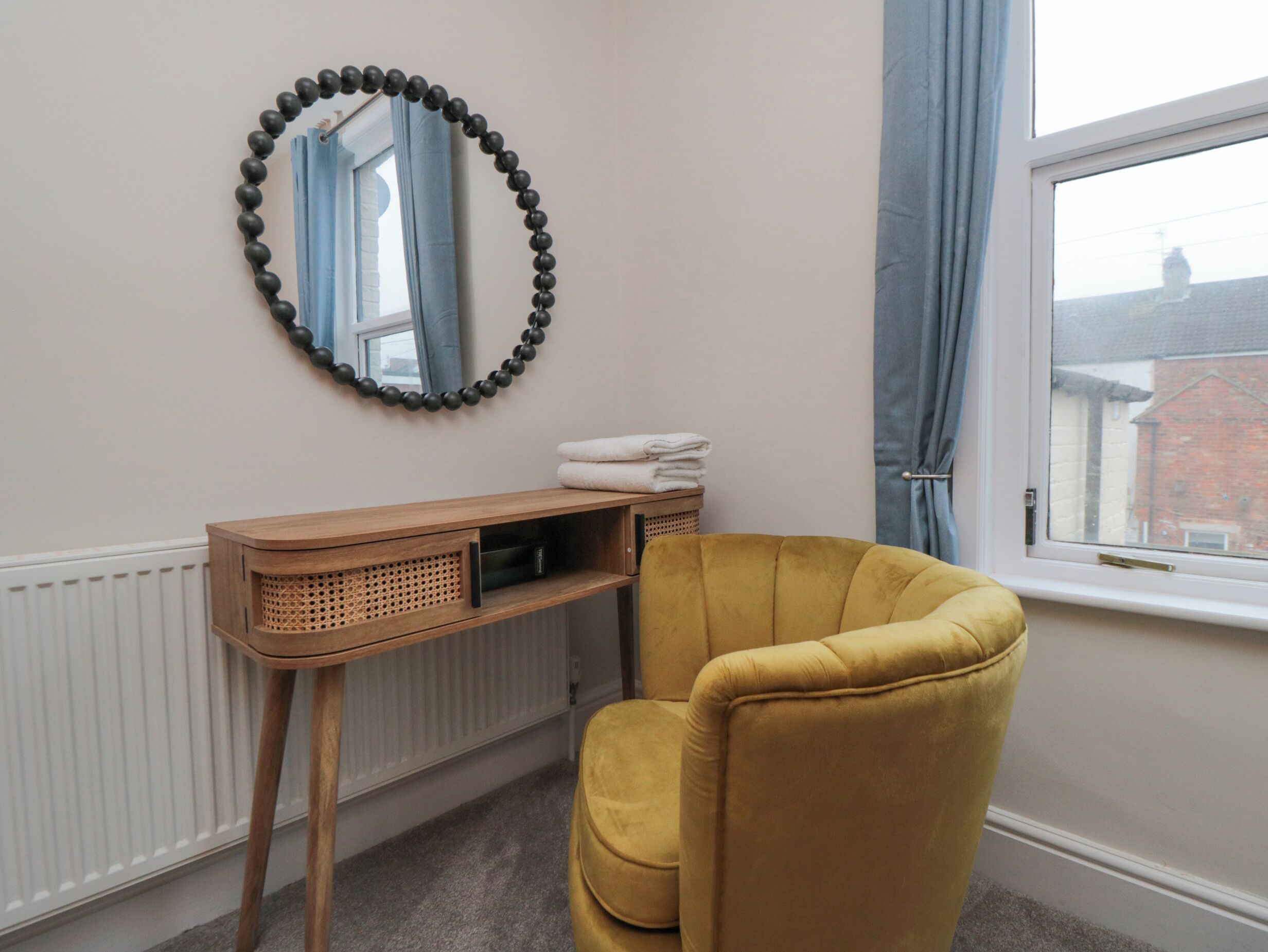 A room with a yellow armchair, a wooden console table with a mirror, and a window with curtains.