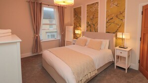 Ein Schlafzimmer mit einem großen Bett, zwei Nachttischen, einer Chandelierleuchtre und einem Fenster mit Vorhängen, an den Wänden blumenmusterter Wandteppich.