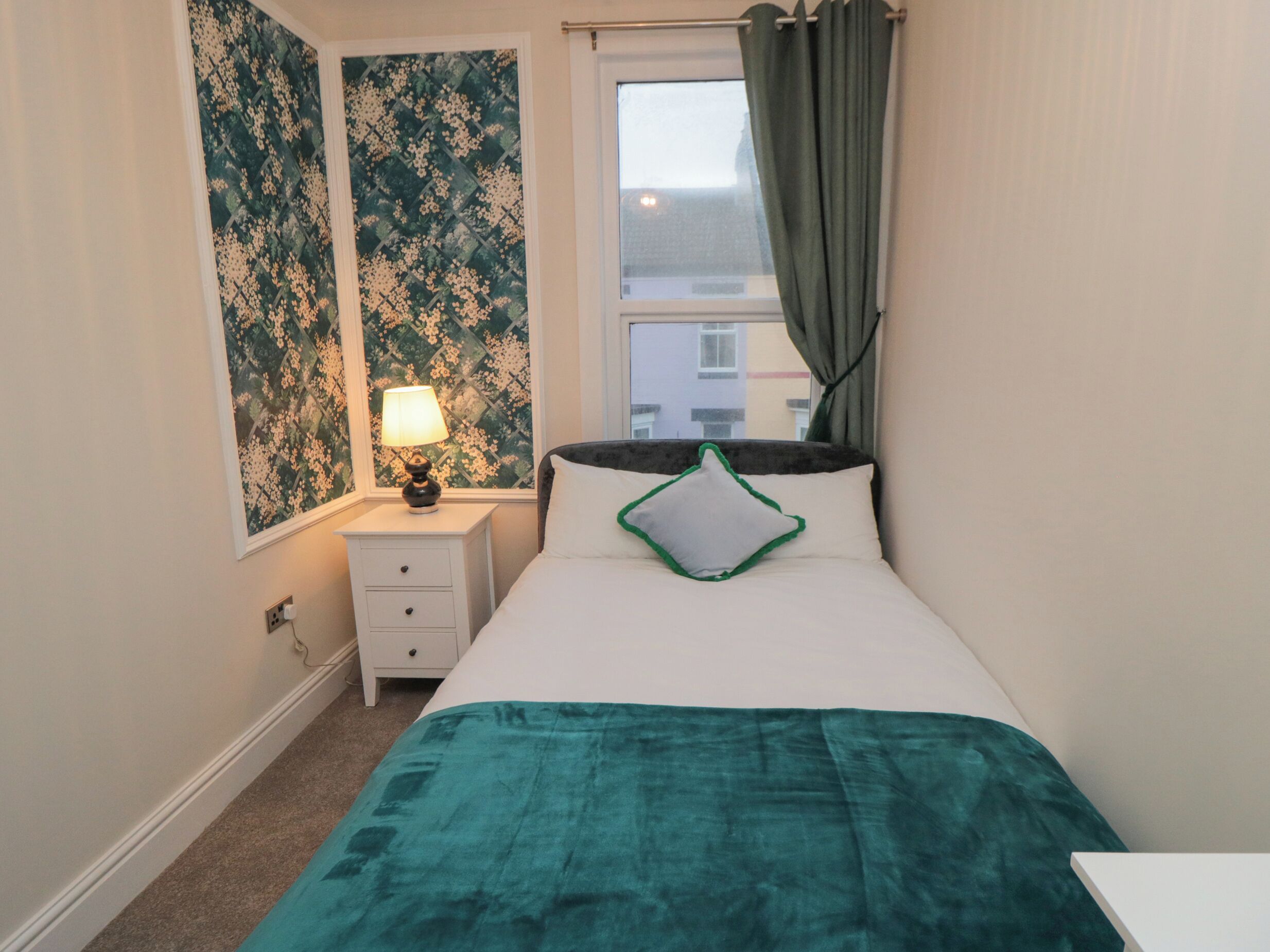 A bedroom with a bed, a nightstand, a lamp, a window with curtains, and a wall with floral wallpaper.