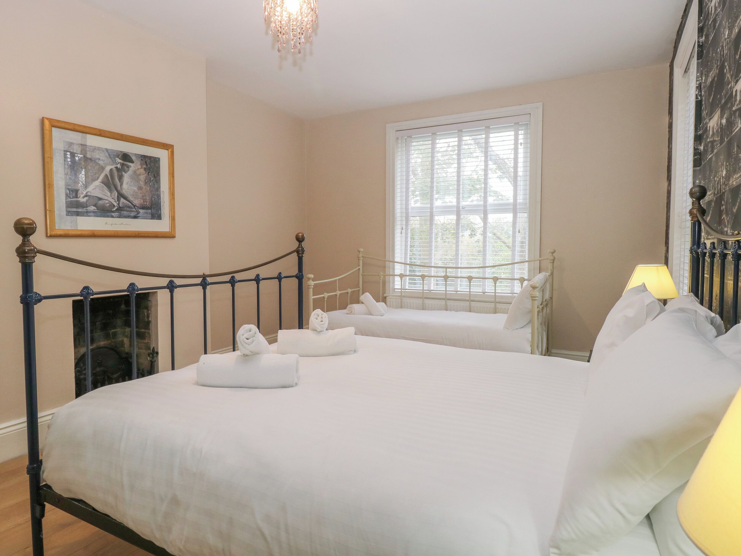 A neatly made double bed with white linens, a bedside table with a lamp, and a window with blinds.