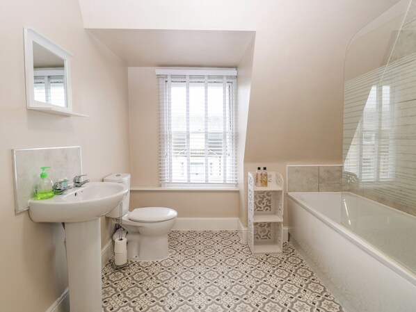A bathroom with a toilet, sink, and bathtub. There is a window with blinds, a mirror, and a shelf with toiletries.