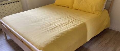 A bed with a yellow bedspread and pillows, a wooden headboard, and a radiator in the background.