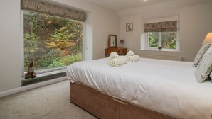 Une chambre avec une grande fenêtre, un lit avec des serviettes pliées, une commode et une table de chevet avec une lampe.