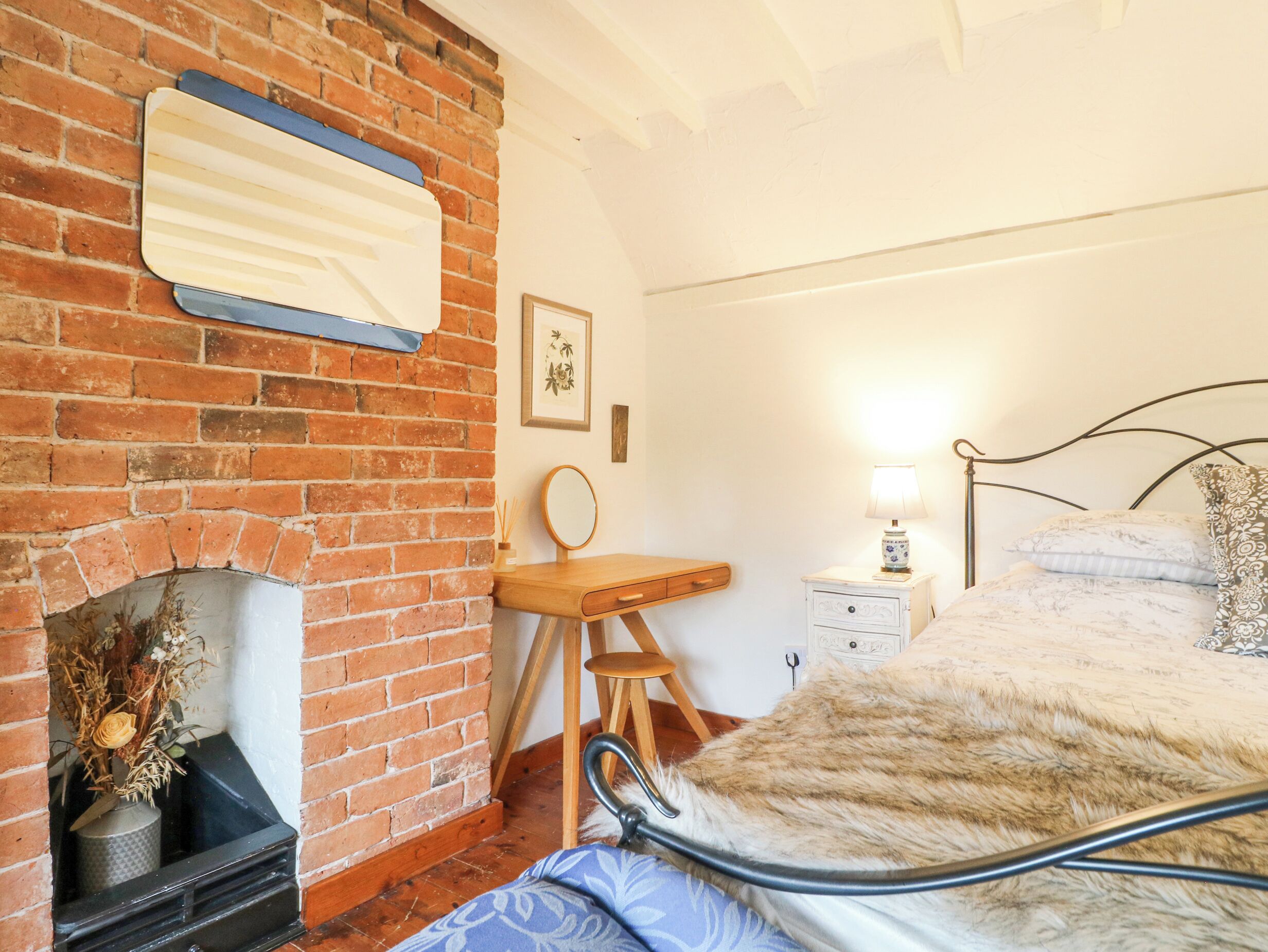 Une chambre avec une cheminée en briques, un bureau en bois, une table de chevet avec une lampe et un lit recouvert d’un plaid en fourrure.