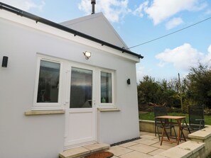 A white building with a flat roof, a door, and two windows. There is a patio area with a table and chairs.