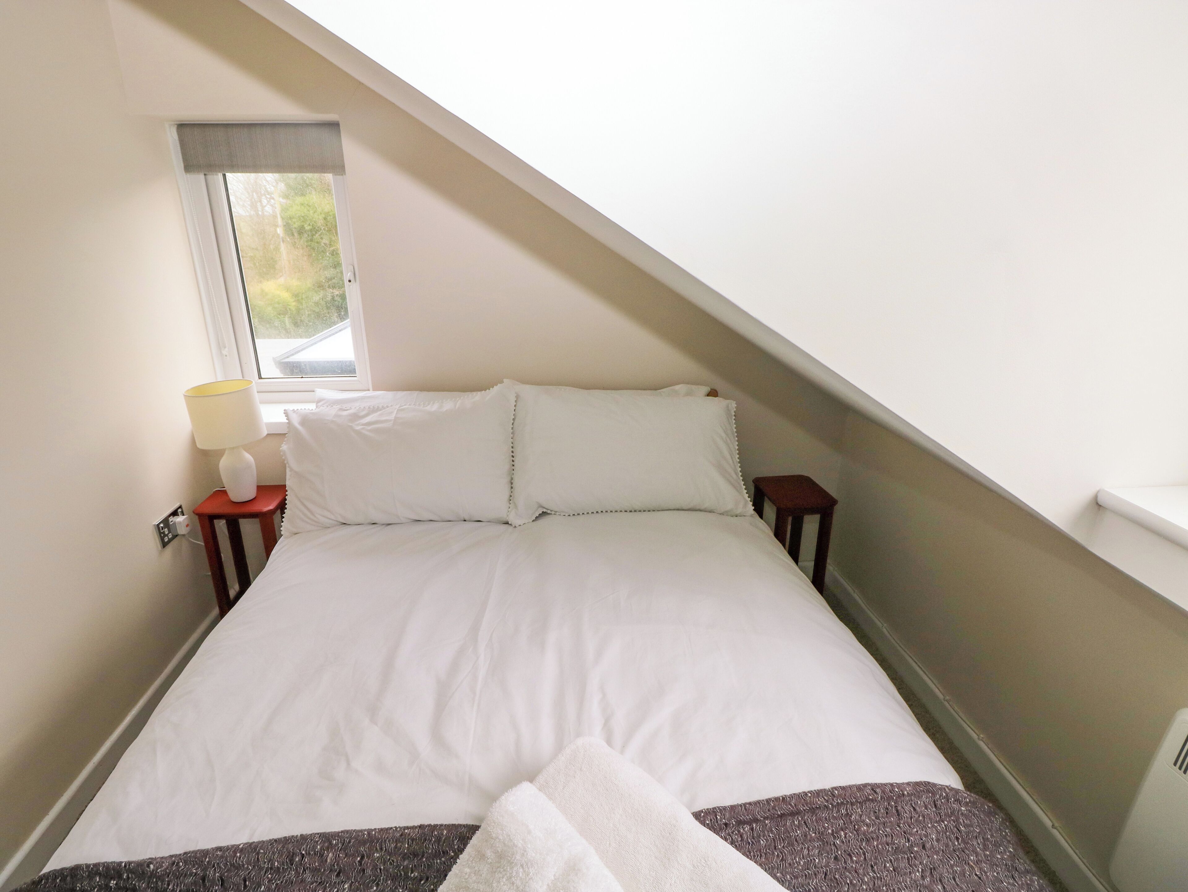 Un dormitorio con cama, una mesita de noche con lámpara, una ventana y vistas al exterior con vegetación.