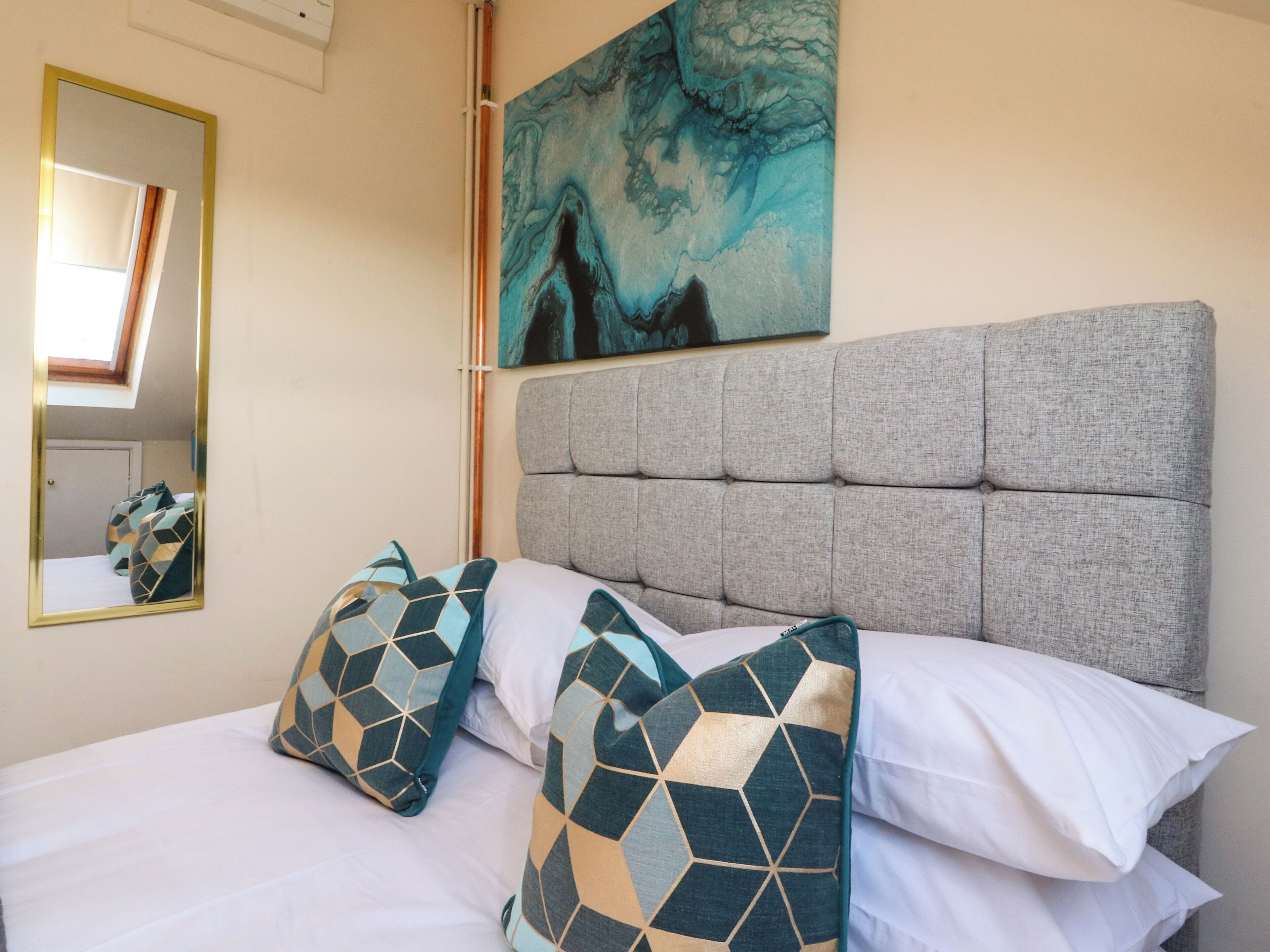 A neatly made bed with patterned pillows, a headboard, a painting on the wall, and a mirror.